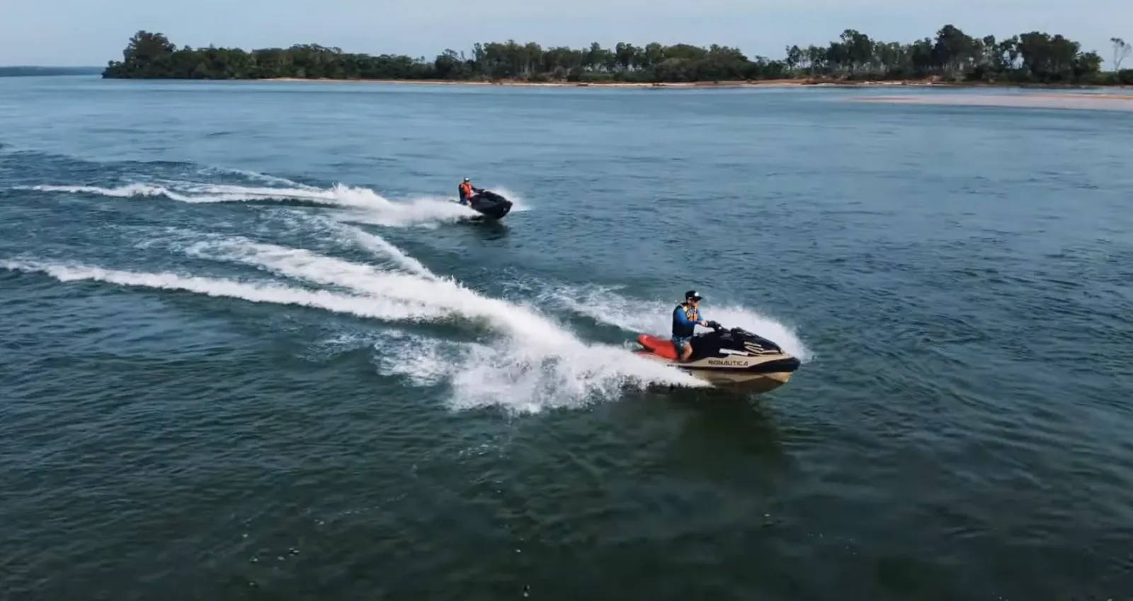 Dois motonautas em motos aquáticas separadas, navegando em alta velocidade pelo Rio Paraná, deixando rastros brancos na água escura, com vegetação rasteira nas margens