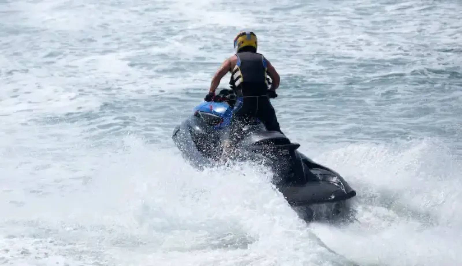 Motonauta visto de costas, usando capacete amarelo e colete salva-vidas, enquanto manobra uma moto aquática preta em alta velocidade, levantando bastante spray