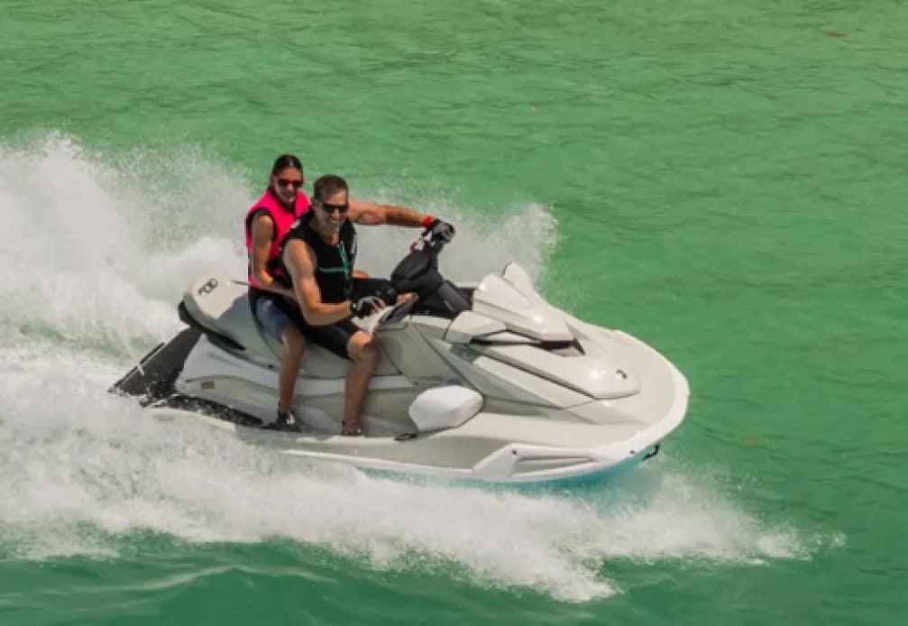 Casal se divertindo em uma moto aquática branca, com o homem pilotando e a mulher na garupa, enquanto fazem uma curva em águas verde-claras