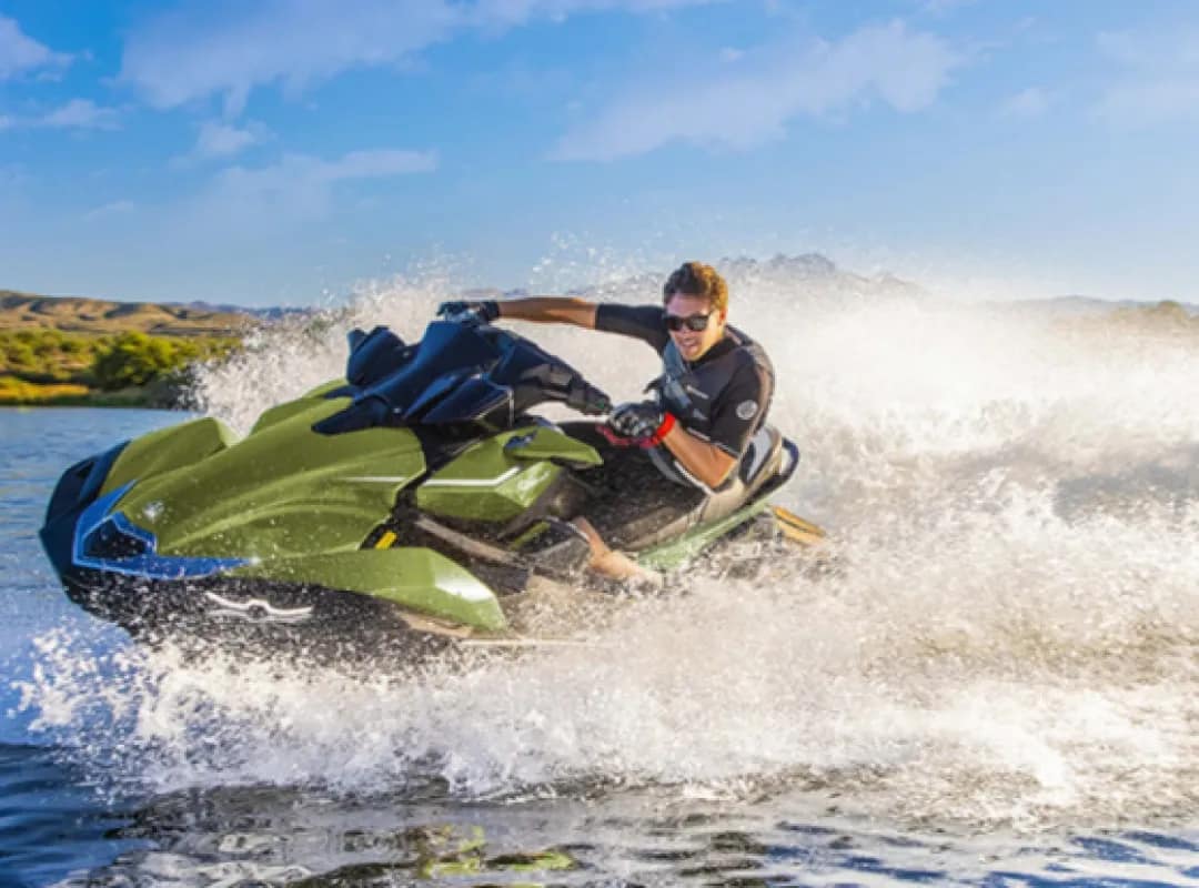 Homem usando o modelo de Jet Ski Kawasaki ULTRA 310LX. Ele está fazendo manobras radicais. O céu está azul e limpo, a água é escura, e atrás se vê alguns bancos de grama.